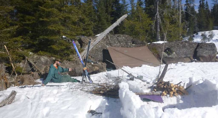 A person rests on the ground at their campsite in the snow. There is a small tarp shelter, firewood and skis. 